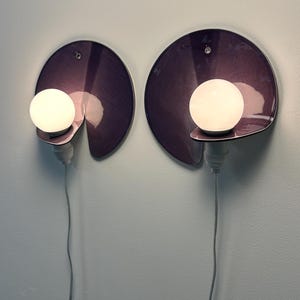 K&ouml;nnte beinhalten: Zwei Wandleuchten mit kreisf&ouml;rmigen, tiefvioletten, gl&auml;nzenden R&uuml;ckplatten mit einer Aussparung. Jede Lampe hat eine wei&szlig;e, kugelf&ouml;rmige Gl&uuml;hbirne auf einer kleinen hervorstehenden Ablage, die mit einem vertikal h&auml;ngenden wei&szlig;en Kabel verbunden ist.