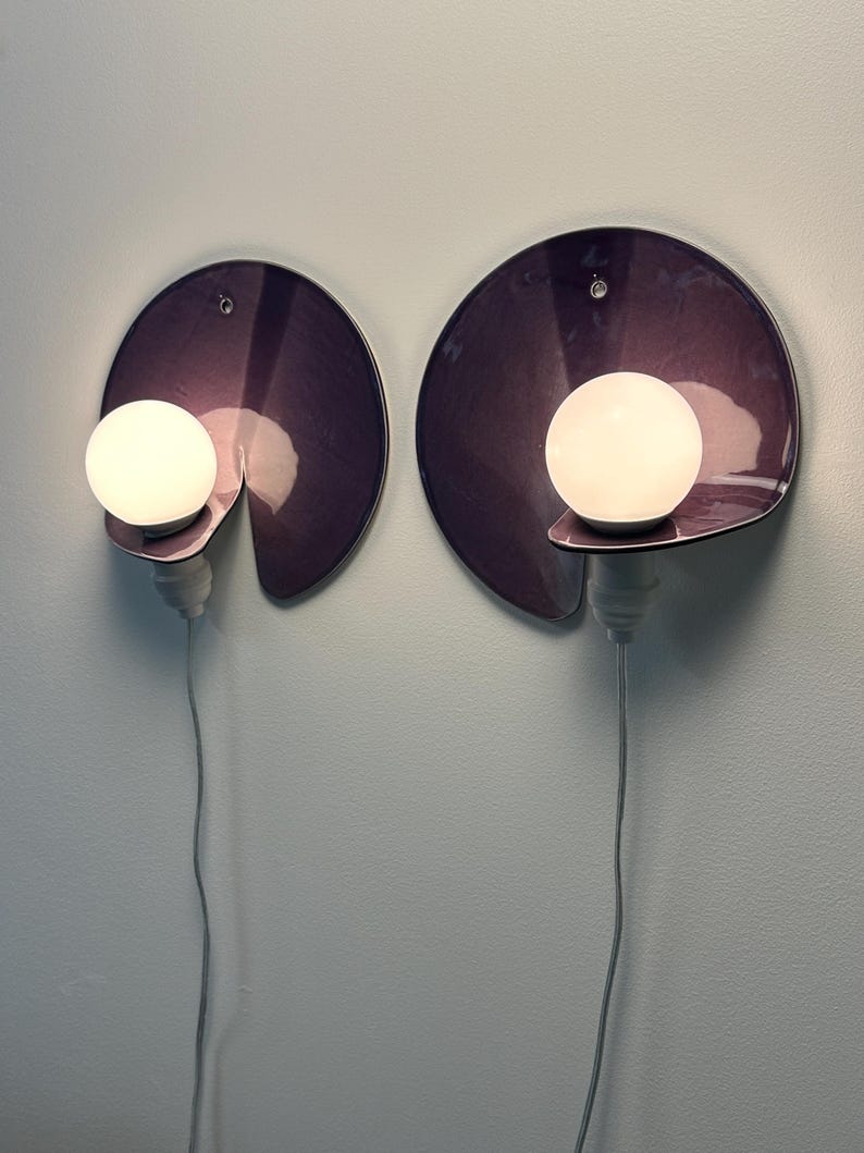 K&ouml;nnte beinhalten: Zwei Wandleuchten mit kreisf&ouml;rmigen, tiefvioletten, gl&auml;nzenden R&uuml;ckplatten mit einer Aussparung. Jede Lampe hat eine wei&szlig;e, kugelf&ouml;rmige Gl&uuml;hbirne auf einer kleinen hervorstehenden Ablage, die mit einem vertikal h&auml;ngenden wei&szlig;en Kabel verbunden ist.
