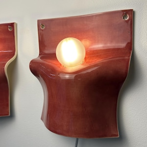 May include: A wall-mounted sconce featuring a glossy, deep red ceramic body with a curved, organic shape. A single, round, frosted light bulb is centered in the middle of the fixture. The ceramic piece has two small holes at the top for mounting.