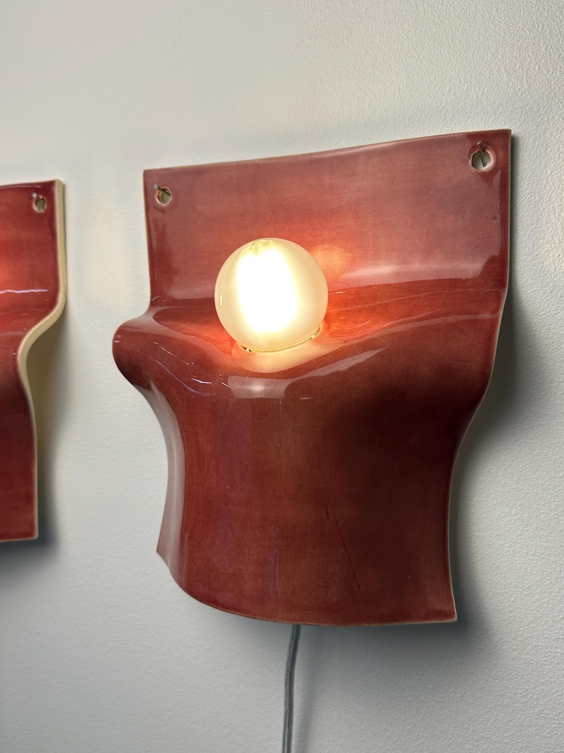 May include: A wall-mounted sconce featuring a glossy, deep red ceramic body with a curved, organic shape. A single, round, frosted light bulb is centered in the middle of the fixture. The ceramic piece has two small holes at the top for mounting.