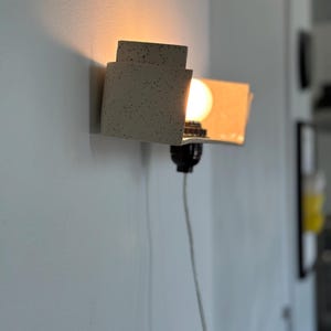 May include: A modern wall sconce with a speckled, off-white finish. The sconce has a layered, geometric design with a warm light bulb. A black socket and a white cord are visible. The sconce is mounted on a light gray wall.