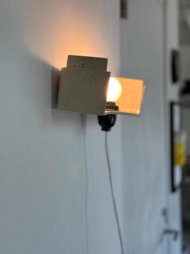 May include: A modern wall sconce with a speckled, off-white finish. The sconce has a layered, geometric design with a warm light bulb. A black socket and a white cord are visible. The sconce is mounted on a light gray wall.