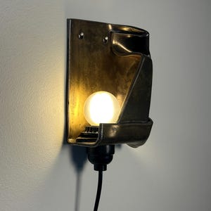 May include: A wall-mounted sconce featuring a dark, metallic, folded rectangular shade with a warm, glowing spherical light bulb. The fixture includes a black socket base and a black power cord extending downward from the bottom of the unit.