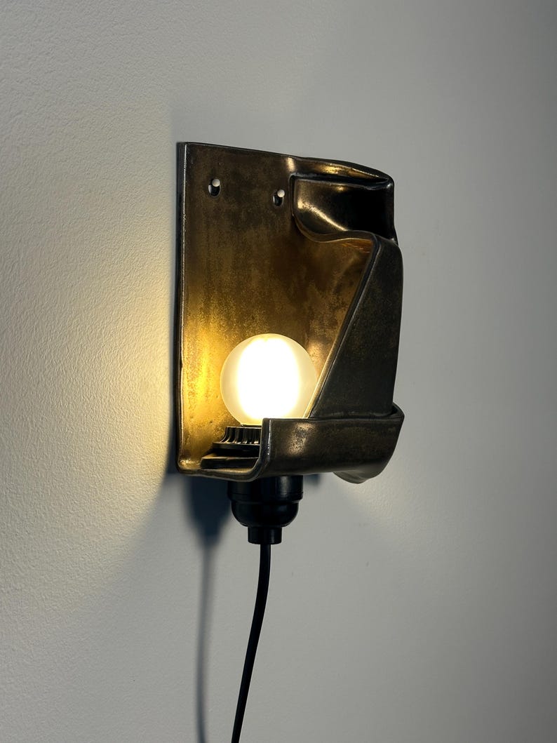 May include: A wall-mounted sconce featuring a dark, metallic, folded rectangular shade with a warm, glowing spherical light bulb. The fixture includes a black socket base and a black power cord extending downward from the bottom of the unit.