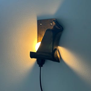 May include: A wall-mounted lamp featuring a dark, curved metal shade that partially obscures a glowing light bulb. The fixture has a rectangular backplate with two visible mounting holes and a black power cord extending from the bottom socket.