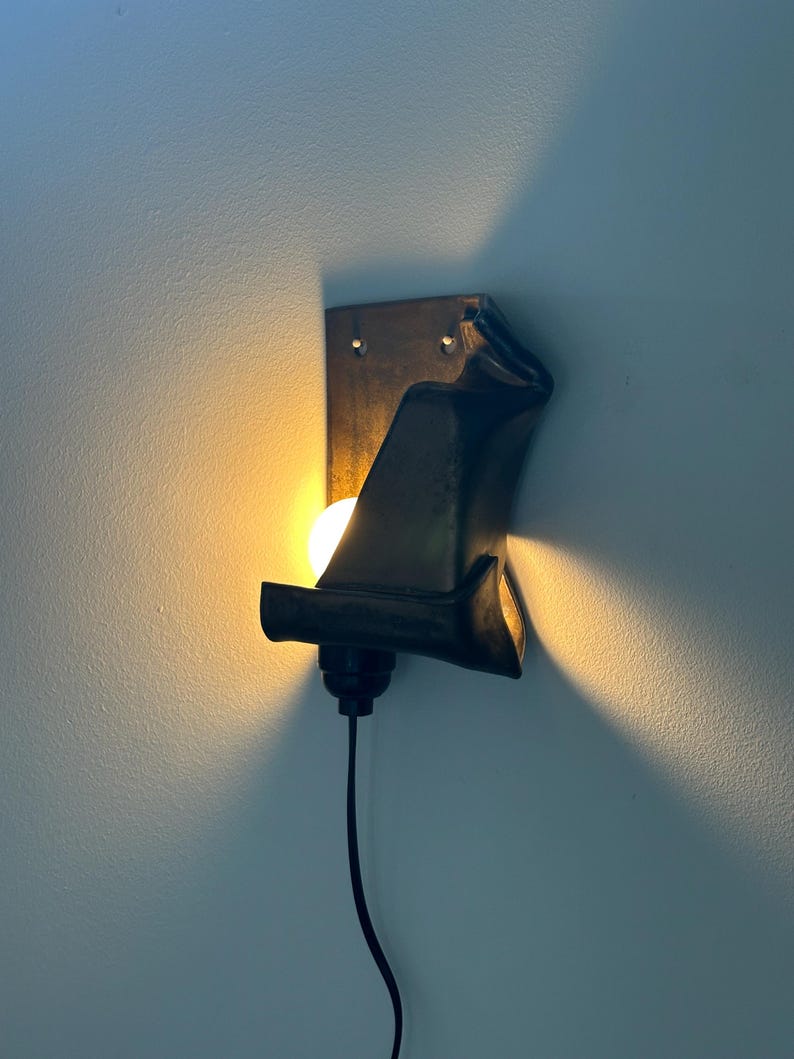 May include: A wall-mounted lamp featuring a dark, curved metal shade that partially obscures a glowing light bulb. The fixture has a rectangular backplate with two visible mounting holes and a black power cord extending from the bottom socket.