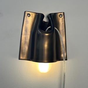 May include: A dark bronze-colored wall sconce with a curved, folded metal design. The fixture features two circular mounting holes at the top, a central opening for a clear power cord, and a single glowing light bulb extending from the bottom.