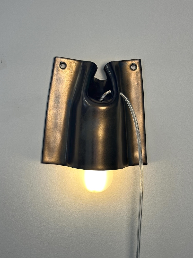 May include: A dark bronze-colored wall sconce with a curved, folded metal design. The fixture features two circular mounting holes at the top, a central opening for a clear power cord, and a single glowing light bulb extending from the bottom.