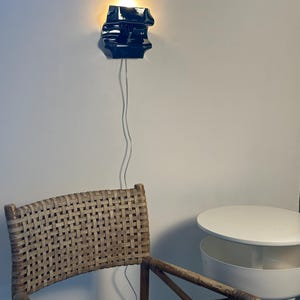 May include: A glossy black ceramic wall sconce with an irregular, sculptural form emits a warm, glowing light. The fixture is mounted on a wall and features a thin, transparent power cord extending downward toward a woven rattan chair.