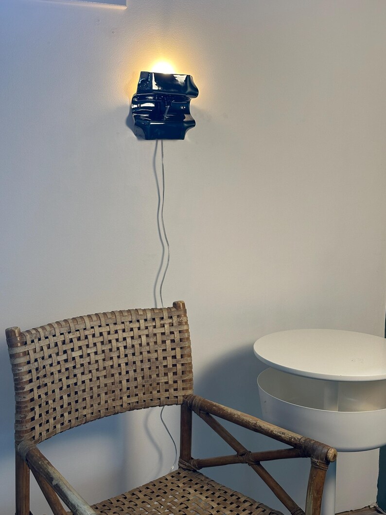 May include: A glossy black ceramic wall sconce with an irregular, sculptural form emits a warm, glowing light. The fixture is mounted on a wall and features a thin, transparent power cord extending downward toward a woven rattan chair.