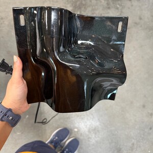 Puede incluir: Un componente de pl&aacute;stico negro brillante moldeado con una superficie irregular y ondulada. La pieza presenta dos ranuras de montaje rectangulares en las esquinas superiores y una peque&ntilde;a abertura circular en el centro, con un cable el&eacute;ctrico.