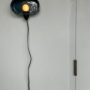May include: A teal ceramic wall sconce with a warm-toned bulb. The sconce has a textured surface and a black cord. The light fixture is mounted on a white wall.