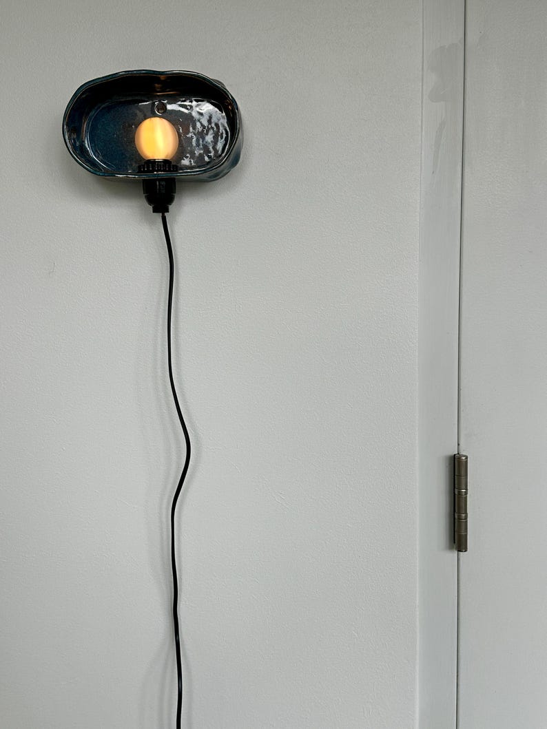 May include: A teal ceramic wall sconce with a warm-toned bulb. The sconce has a textured surface and a black cord. The light fixture is mounted on a white wall.