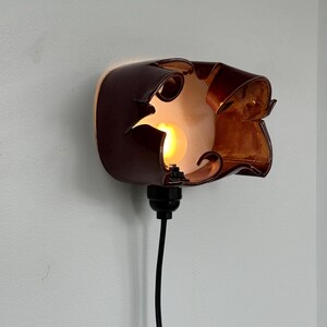 May include: A unique, brown wall sconce with a warm, amber glow. The fixture has an organic, sculpted design with cut-out details. A black cord extends from the base, ready for plugging in. The light bulb is visible through the openings.