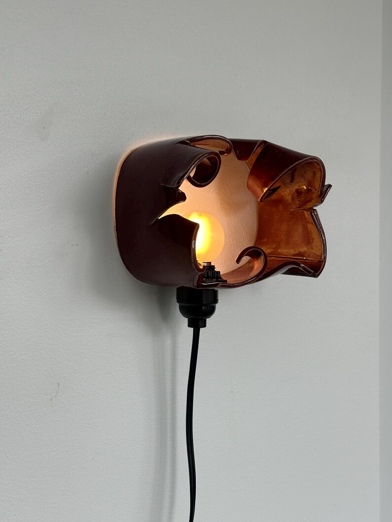 May include: A unique, brown wall sconce with a warm, amber glow. The fixture has an organic, sculpted design with cut-out details. A black cord extends from the base, ready for plugging in. The light bulb is visible through the openings.
