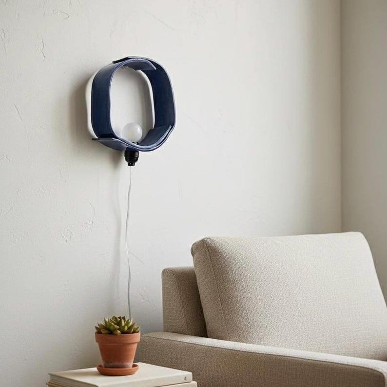 Puede incluir: Aplique de pared moderno, azul marino, con interior blanco, con forma de letra O estilizada. La l&aacute;mpara tiene una bombilla y una base negra, con un cable blanco. El aplique est&aacute; montado en una pared blanca.