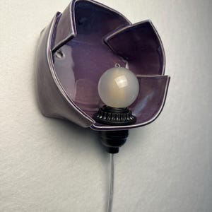 K&ouml;nnte beinhalten: Eine Wandleuchte mit einem gl&auml;nzenden, tiefvioletten Keramikschirm in Form einer offenen Bl&uuml;te. Eine kleine, runde, mattwei&szlig;e Gl&uuml;hbirne befindet sich in der Mitte der Keramikbl&auml;tter und ist an einem schwarzen Sockel mit transparentem Kabel befestigt.