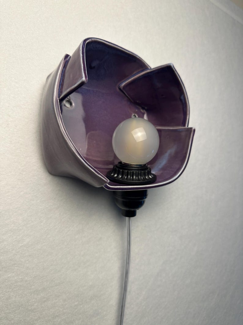K&ouml;nnte beinhalten: Eine Wandleuchte mit einem gl&auml;nzenden, tiefvioletten Keramikschirm in Form einer offenen Bl&uuml;te. Eine kleine, runde, mattwei&szlig;e Gl&uuml;hbirne befindet sich in der Mitte der Keramikbl&auml;tter und ist an einem schwarzen Sockel mit transparentem Kabel befestigt.