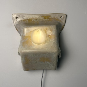 May include: A wall-mounted light fixture featuring a textured, off-white and yellow mottled ceramic housing with a flared top edge and two mounting holes. A single, round, glowing light bulb is centered on the front face, with a thin cord extending.