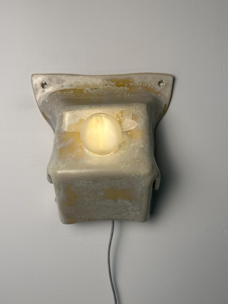 May include: A wall-mounted light fixture featuring a textured, off-white and yellow mottled ceramic housing with a flared top edge and two mounting holes. A single, round, glowing light bulb is centered on the front face, with a thin cord extending.