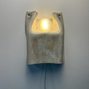 May include: A wall-mounted ceramic wall light with a textured, off-white glaze and a central cutout. A glowing, frosted glass orb bulb is nestled within the upper opening of the fixture. A thin, light-coloured electrical cord extends from the bottom.