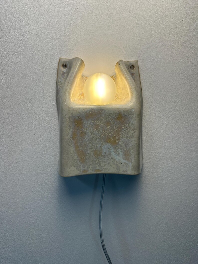 May include: A wall-mounted ceramic wall light with a textured, off-white glaze and a central cutout. A glowing, frosted glass orb bulb is nestled within the upper opening of the fixture. A thin, light-coloured electrical cord extends from the bottom.