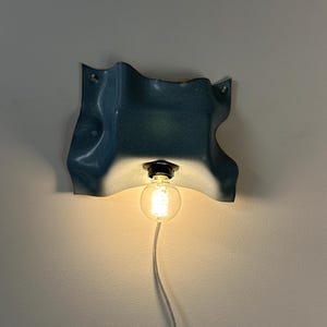 May include: A wall-mounted light fixture featuring a wavy, blue-toned ceramic shade with a glossy finish. A single round, glowing light bulb is suspended from the center of the shade, connected to a white power cord hanging downward.