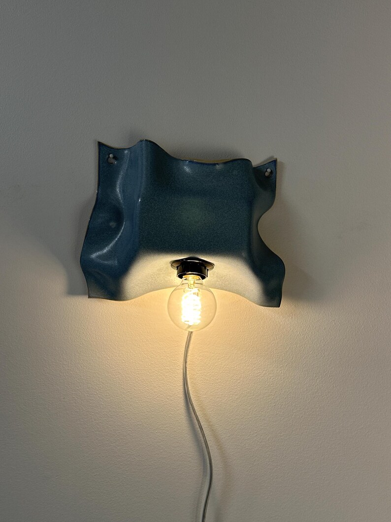 May include: A wall-mounted light fixture featuring a wavy, blue-toned ceramic shade with a glossy finish. A single round, glowing light bulb is suspended from the center of the shade, connected to a white power cord hanging downward.