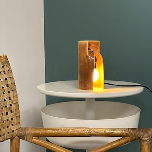 Puede incluir: Una l&aacute;mpara de mesa cil&iacute;ndrica con un acabado de resina en tonos &aacute;mbar y marr&oacute;n moteado. La l&aacute;mpara presenta una secci&oacute;n recortada vertical que revela una bombilla encendida en su interior, conectada a un cable de alimentaci&oacute;n negro.
