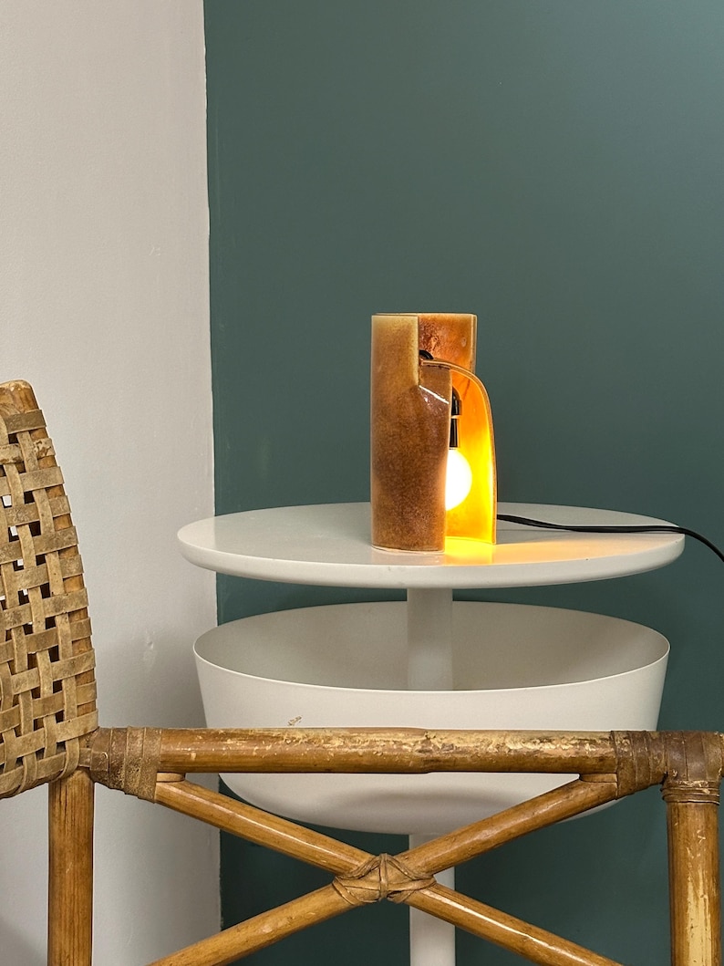 Puede incluir: Una l&aacute;mpara de mesa cil&iacute;ndrica con un acabado de resina en tonos &aacute;mbar y marr&oacute;n moteado. La l&aacute;mpara presenta una secci&oacute;n recortada vertical que revela una bombilla encendida en su interior, conectada a un cable de alimentaci&oacute;n negro.