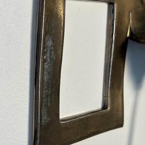 K&ouml;nnte beinhalten: Ein metallischer, bronzefarbener Rahmen mit einem rechteckigen Ausschnitt. Die Oberfl&auml;che weist ein verwittertes, strukturiertes Finish auf, mit dunkleren Schattierungen an den Kanten und einem helleren, leicht abgenutzten Aussehen.