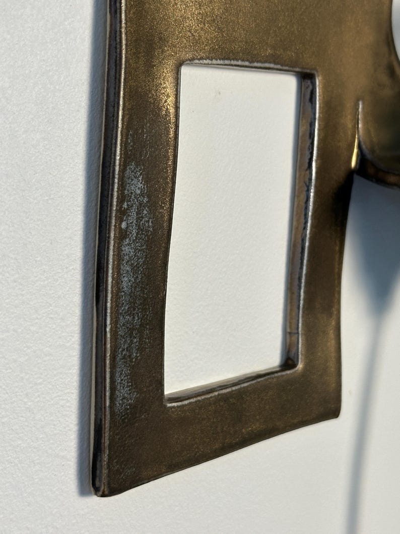 K&ouml;nnte beinhalten: Ein metallischer, bronzefarbener Rahmen mit einem rechteckigen Ausschnitt. Die Oberfl&auml;che weist ein verwittertes, strukturiertes Finish auf, mit dunkleren Schattierungen an den Kanten und einem helleren, leicht abgenutzten Aussehen.