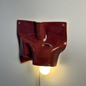 May include: A wall-mounted ceramic light fixture with a glossy, deep red finish and an irregular, folded shape. The piece features two circular mounting holes at the top corners, a small silver metal ring detail, and a single glowing light bulb.
