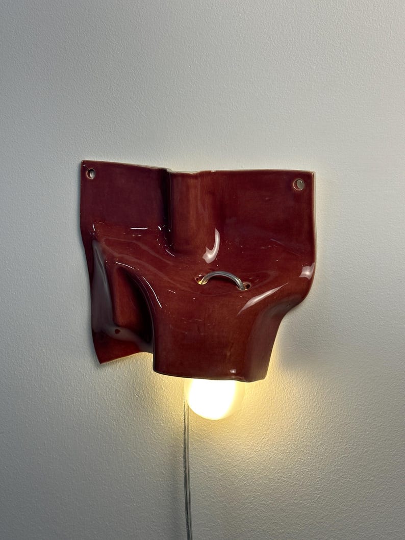 May include: A wall-mounted ceramic light fixture with a glossy, deep red finish and an irregular, folded shape. The piece features two circular mounting holes at the top corners, a small silver metal ring detail, and a single glowing light bulb.