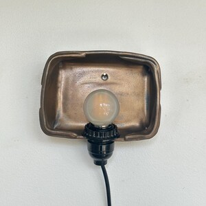 May include: A bronze-colored wall sconce with a rectangular backplate. A black light socket holds a round, clear light bulb. The sconce is mounted on a white wall, and a black cord extends downward.