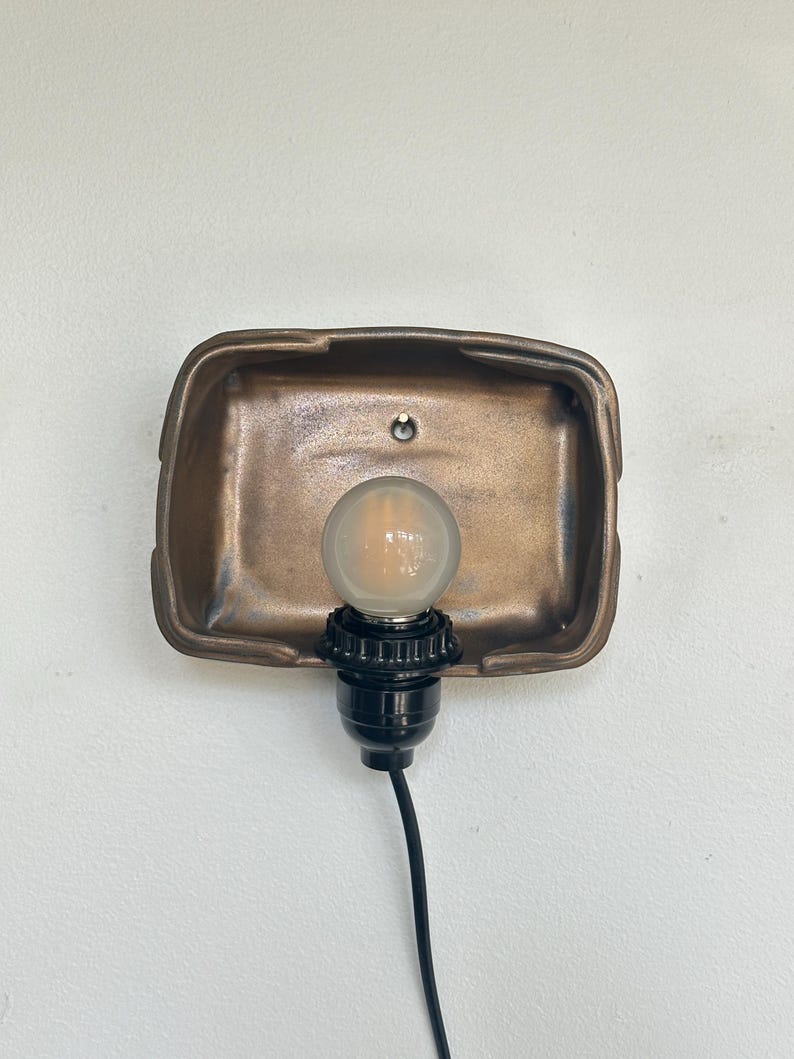 May include: A bronze-colored wall sconce with a rectangular backplate. A black light socket holds a round, clear light bulb. The sconce is mounted on a white wall, and a black cord extends downward.