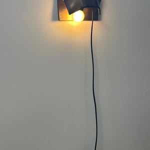 Pode incluir: Uma lumin&aacute;ria de parede com uma c&uacute;pula preta fosca curva que direciona a luz para baixo. Uma &uacute;nica l&acirc;mpada brilhante de tom quente est&aacute; posicionada sob a c&uacute;pula. Um fio de alimenta&ccedil;&atilde;o preto fino desce verticalmente at&eacute; um interruptor.