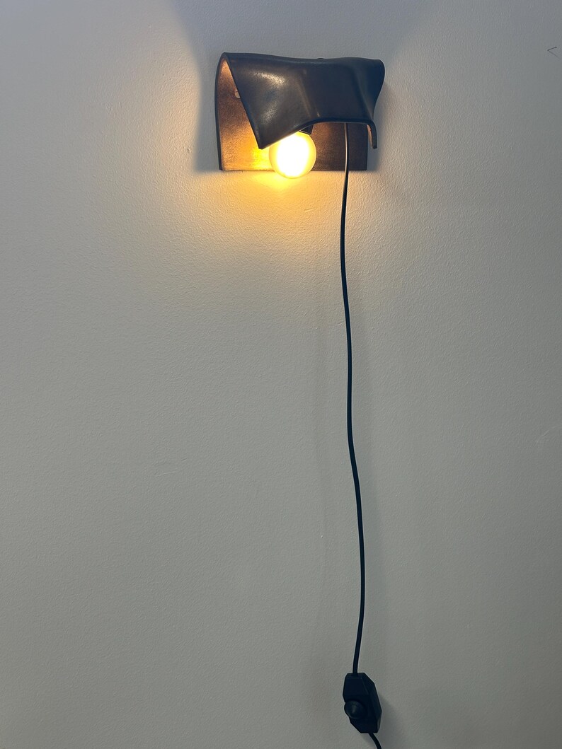 Pode incluir: Uma lumin&aacute;ria de parede com uma c&uacute;pula preta fosca curva que direciona a luz para baixo. Uma &uacute;nica l&acirc;mpada brilhante de tom quente est&aacute; posicionada sob a c&uacute;pula. Um fio de alimenta&ccedil;&atilde;o preto fino desce verticalmente at&eacute; um interruptor.