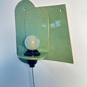 May include: A wall-mounted light fixture featuring a glossy, speckled light green ceramic backplate with an arched design. A black socket holds a round, frosted white light bulb on a small ceramic shelf. A thin power cord hangs from the bottom.