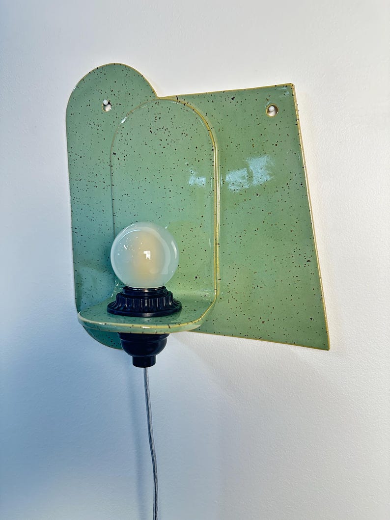 May include: A wall-mounted light fixture featuring a glossy, speckled light green ceramic backplate with an arched design. A black socket holds a round, frosted white light bulb on a small ceramic shelf. A thin power cord hangs from the bottom.