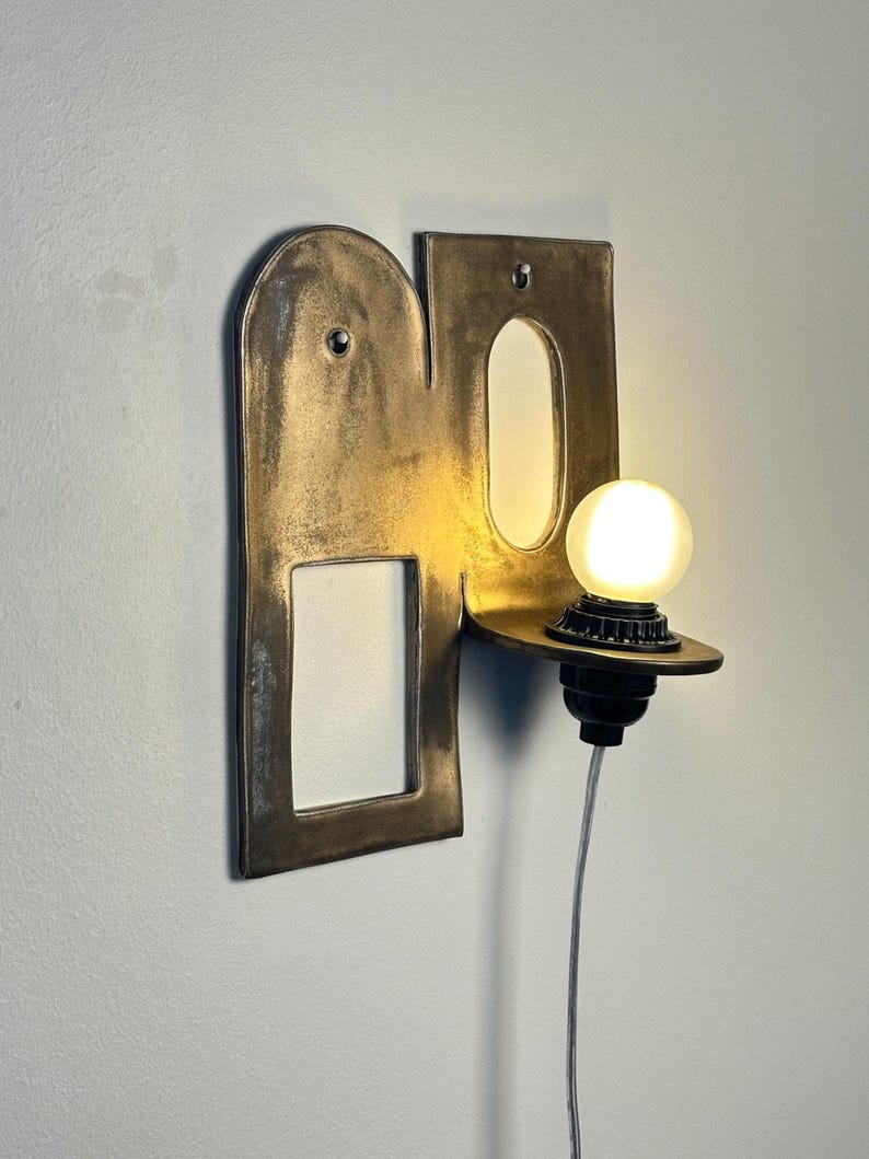 K&ouml;nnte beinhalten: Eine Wandleuchte mit einer metallischen, bronzefarbenen Platte, die einen bogenf&ouml;rmigen Ausschnitt und eine rechteckige &Ouml;ffnung aufweist. Eine kleine, runde, mattierte Gl&uuml;hbirne sitzt auf einer hervorstehenden Ablage, verbunden durch ein d&uuml;nnes, helles Kabel.