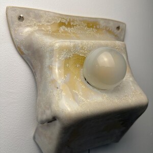 May include: A wall-mounted ceramic light fixture featuring a glossy, cream-colored glaze with yellow and amber crystalline patterns. The square-shaped base has two mounting holes at the top and holds a single, round, frosted white glass light bulb.