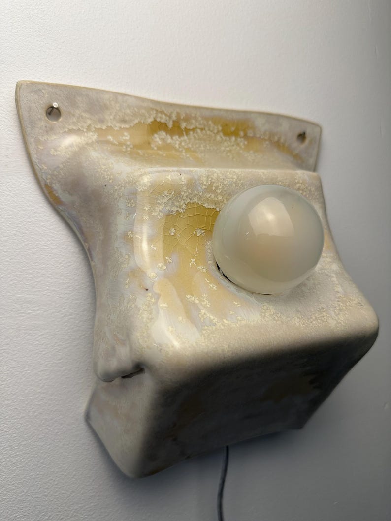 May include: A wall-mounted ceramic light fixture featuring a glossy, cream-colored glaze with yellow and amber crystalline patterns. The square-shaped base has two mounting holes at the top and holds a single, round, frosted white glass light bulb.