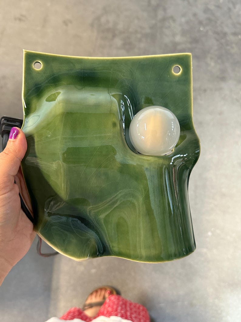 May include: A wall-mounted ceramic lamp with a glossy, deep green glaze and a visible crackle finish. The rectangular form features organic, undulating curves and two circular holes at the top corners. A single round, frosted white light bulb is set into a recessed circular opening on the right side.