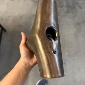 May include: A metallic bronze-toned, tapered, hollow object with a smooth, slightly weathered finish. It features a large circular opening on the side, with a smaller hole below it through which a clear, flexible plastic cord is threaded.