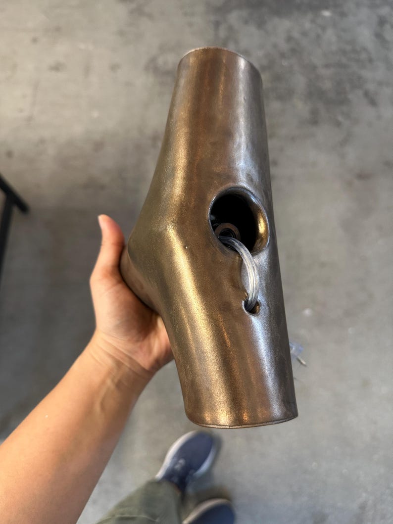 May include: A metallic bronze-toned, tapered, hollow object with a smooth, slightly weathered finish. It features a large circular opening on the side, with a smaller hole below it through which a clear, flexible plastic cord is threaded.