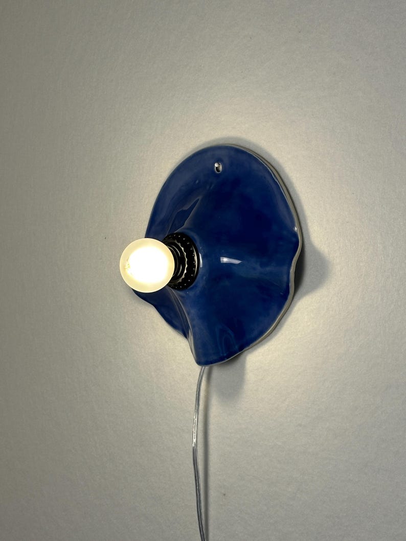 May include: A blue ceramic wall sconce with a bulb. The sconce has a unique, organic shape and a glossy finish. The bulb is lit, emitting a warm glow. A clear cord extends from the sconce.
