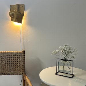 K&ouml;nnte beinhalten: Eine Wandleuchte aus Keramik mit gl&auml;nzend beiger Oberfl&auml;che und geschwungener, organischer Form, die ein warmes Licht ausstrahlt. Daneben steht eine klare, rechteckige Glasvase mit schwarzem Metallrahmen und wei&szlig;en Schleierkrautbl&uuml;ten auf einem runden Tisch.