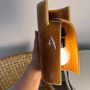 Puede incluir: Un aplique de pared de cer&aacute;mica con un cuerpo cil&iacute;ndrico curvado y un esmalte marr&oacute;n brillante moteado. El dise&ntilde;o abierto revela un casquillo negro que sostiene una bombilla blanca redonda e iluminada, con un cable negro.
