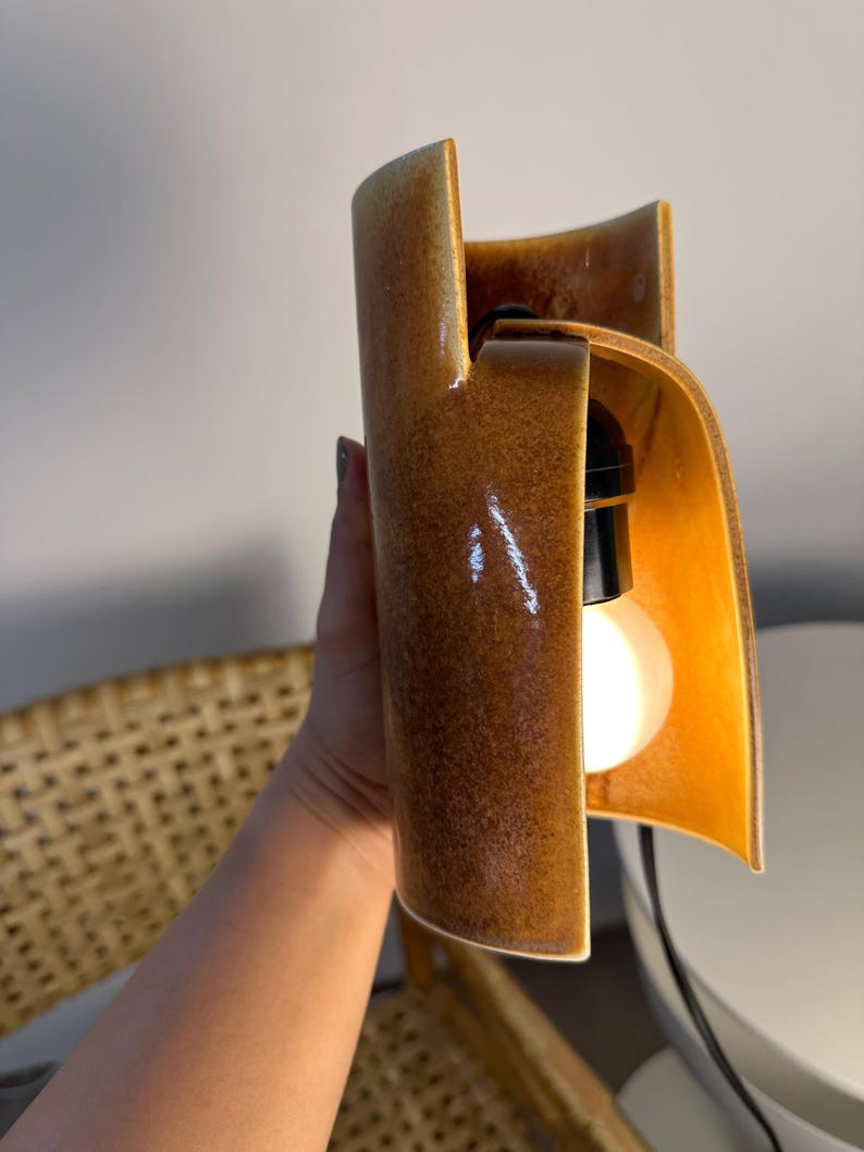 Puede incluir: Un aplique de pared de cer&aacute;mica con un cuerpo cil&iacute;ndrico curvado y un esmalte marr&oacute;n brillante moteado. El dise&ntilde;o abierto revela un casquillo negro que sostiene una bombilla blanca redonda e iluminada, con un cable negro.